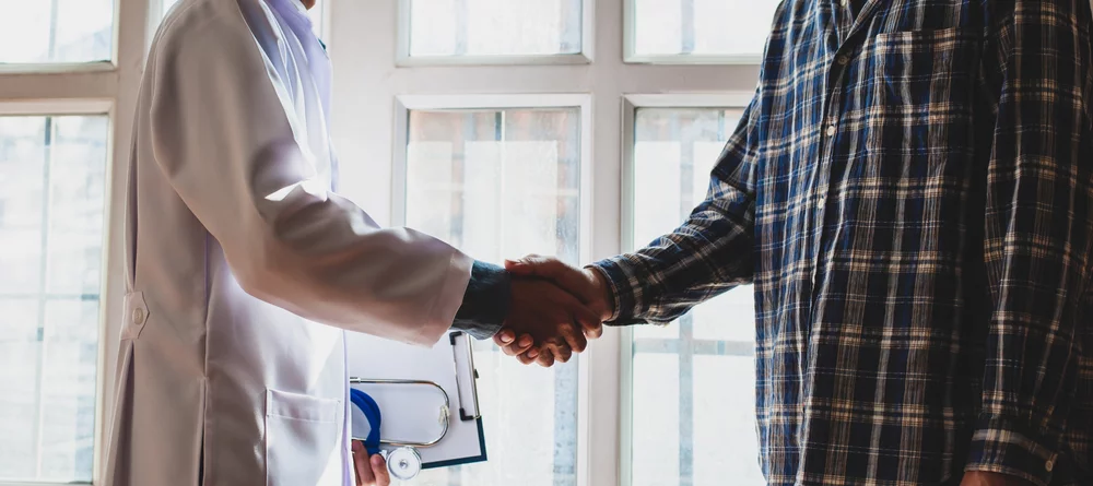A patient shakes a doctor’s hand in a hospital or clinic hallway.